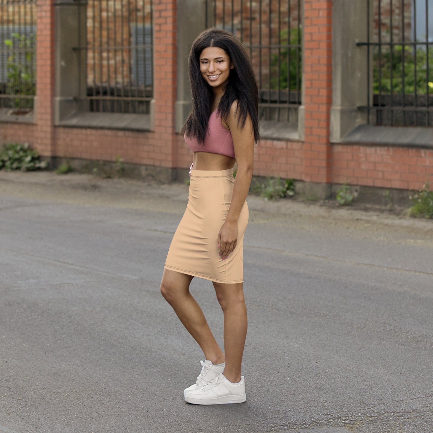 Woman in a pink crop top and beige skirt standing on a street with a brick building in the background.