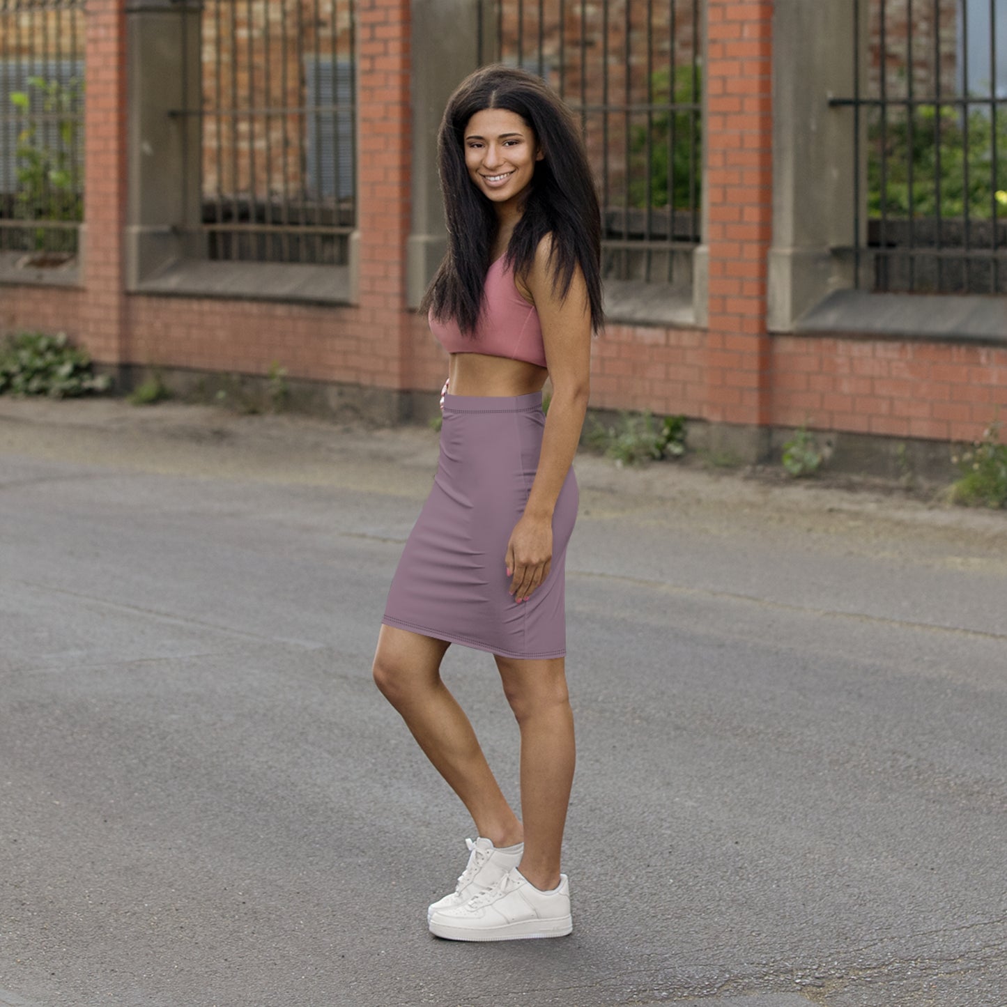 Woman in pink crop top and purple skirt standing on a street with a brick building in the background