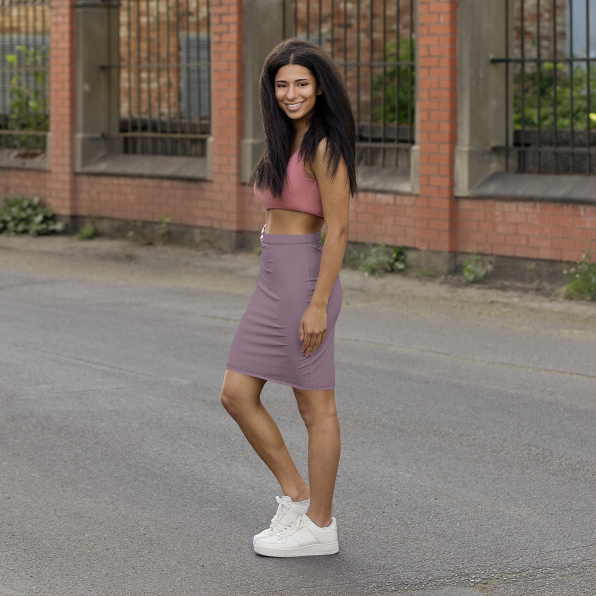 Woman in pink crop top and purple skirt standing on a street with a brick building in the background