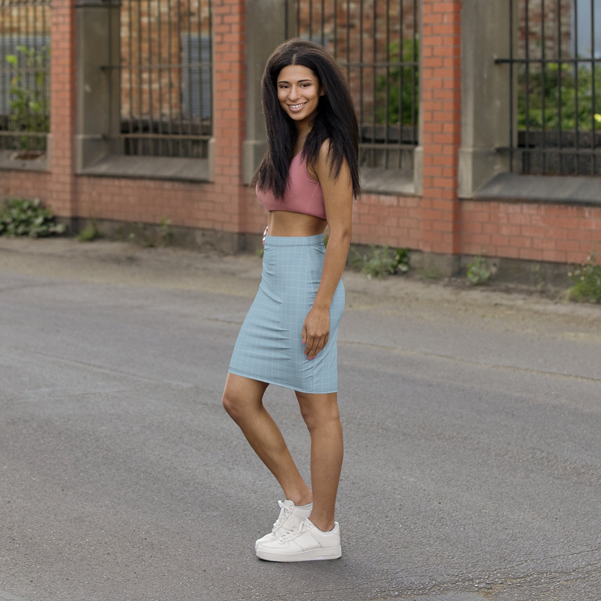 Woman in a pink crop top and light blue skirt standing on a street with a brick building in the background.